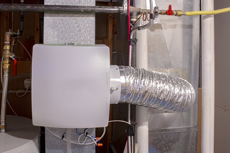 A white whole-house humidifier unit attached to HVAC ductwork with a flexible aluminum bypass tube and surrounding plumbing lines.